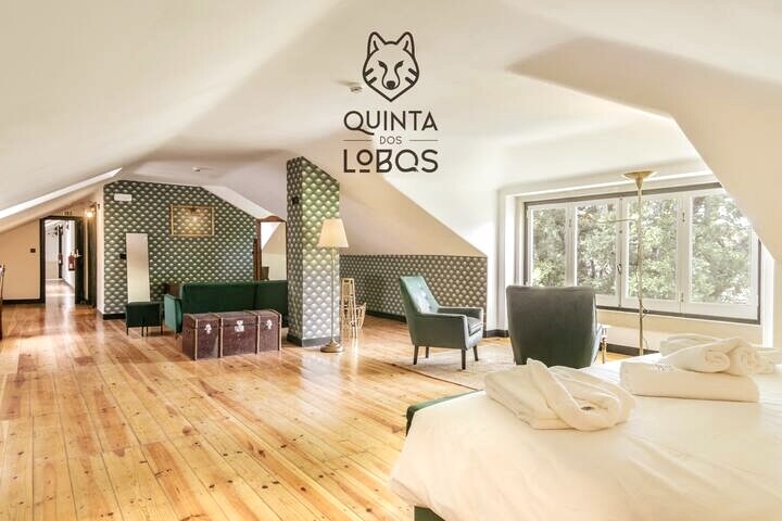 A spacious room is featured with natural wood flooring and large windows allowing ample light, showcasing views of the surrounding greenery. Cozy seating areas are arranged with armchairs and a couch, complemented by modern decor elements. Towels are neatly folded on a spacious bed.
