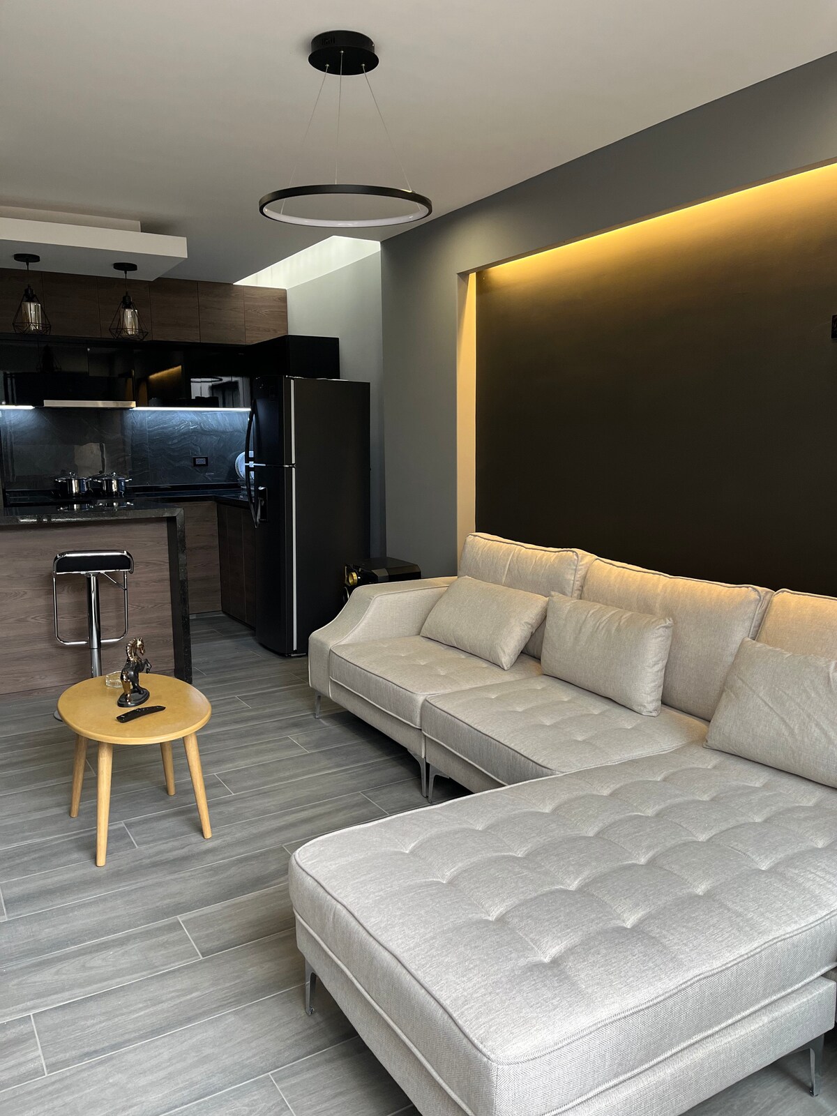 A modern living area features a light gray sectional sofa with tufted detailing, accompanied by a small round table. The kitchen visible in the background includes sleek cabinetry and stainless steel appliances, complemented by ambient lighting that enhances the space's contemporary design.