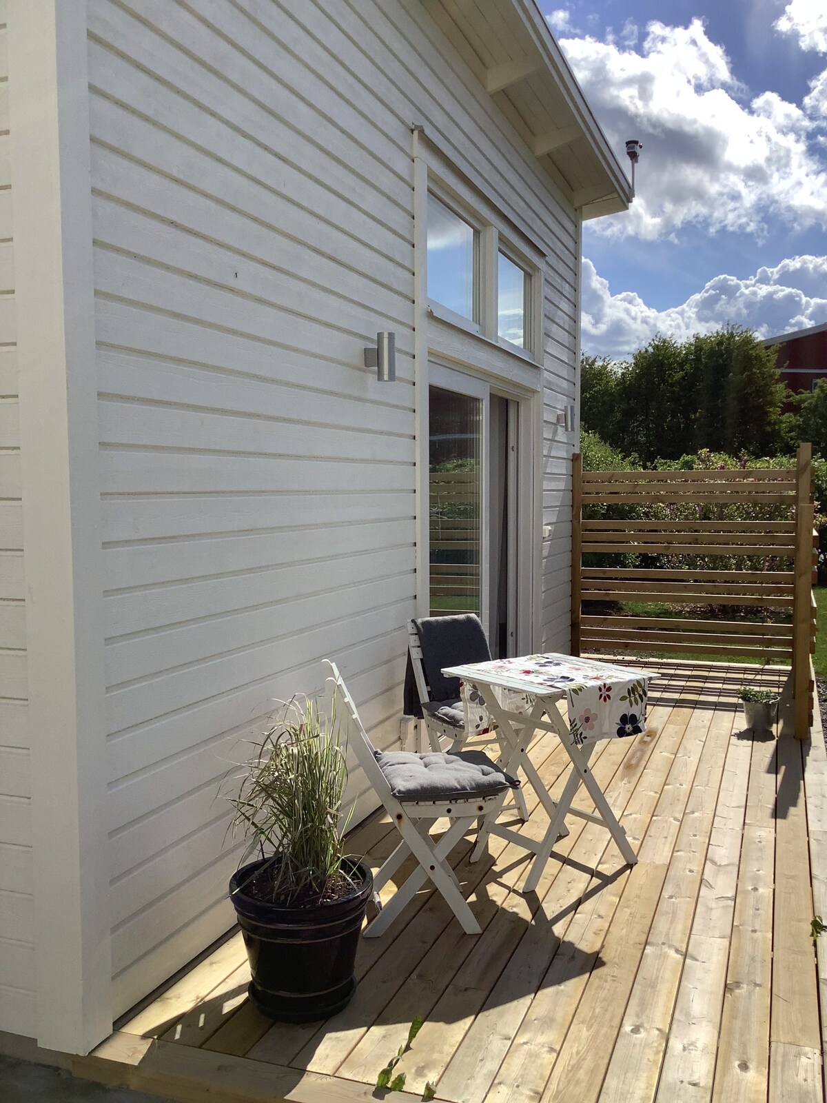 An outdoor patio features a wooden deck with space for a small table and two folding chairs. A potted plant sits beside the table, and a wooden privacy screen partially encloses the area. Sunlight filters through clouds, illuminating the white exterior of the guesthouse.