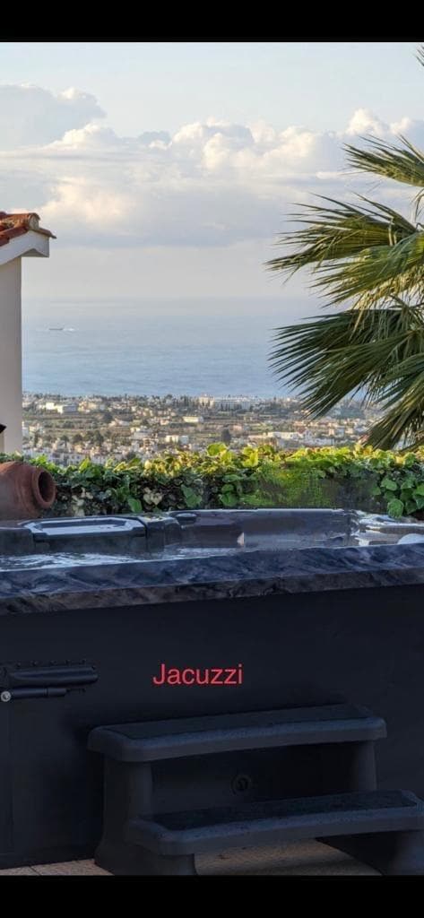 A jacuzzi is positioned with a view of the sea and surrounding landscapes. The area is framed by palm leaves and a terracotta pot, creating a peaceful setting. Steps lead up to the jacuzzi, inviting relaxation against the backdrop of clouds and distant hills.