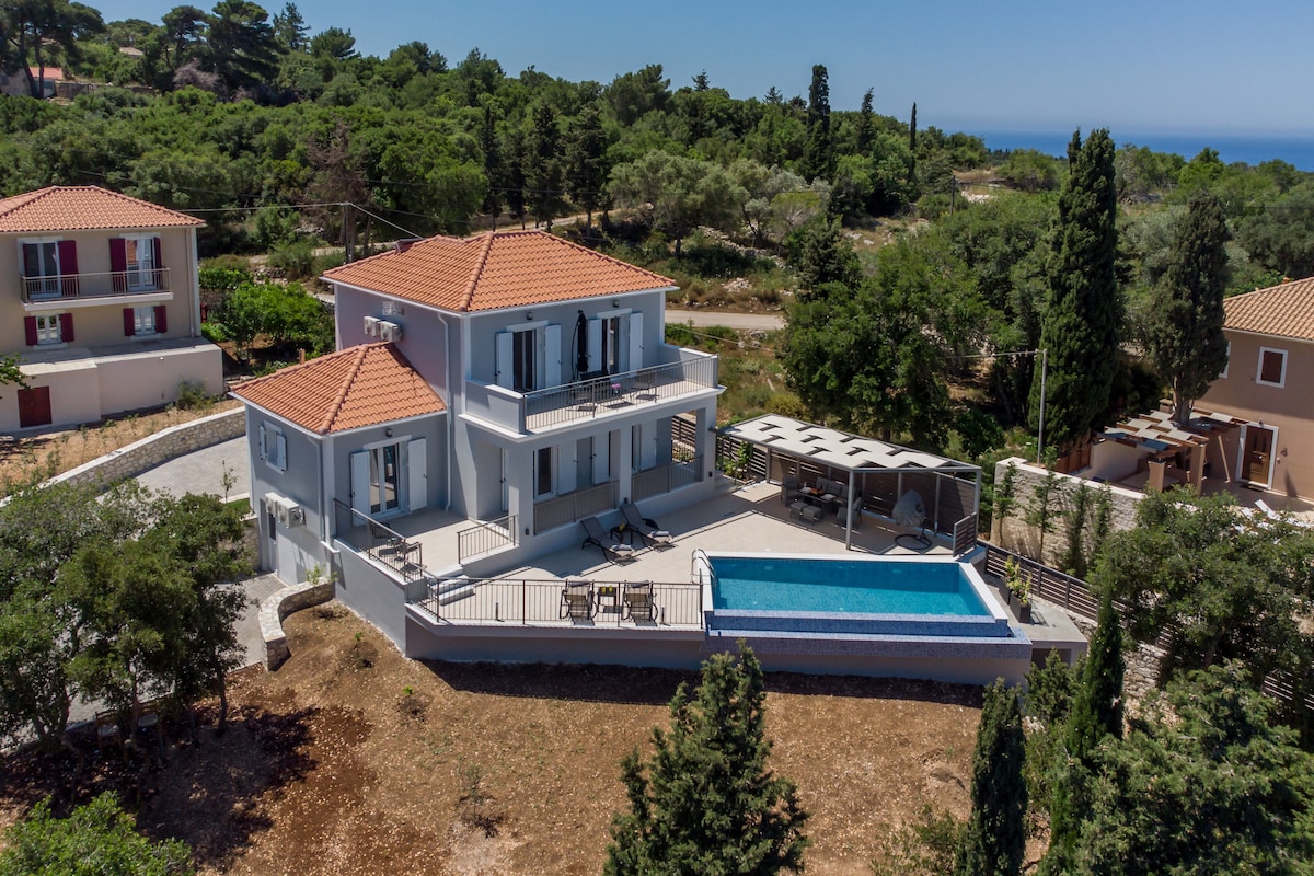 A modern two-story villa is situated amidst lush greenery, featuring a well-maintained outdoor space with a private pool. The villa's design includes balconies with views of the surrounding area, complemented by a shaded terrace. Nearby trees offer additional privacy and tranquility.