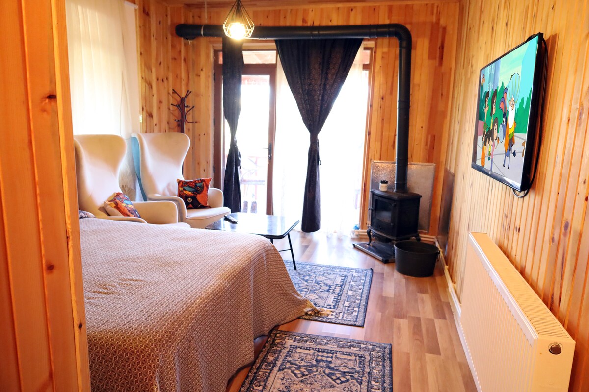 A cozy room features a spacious bed covered with a patterned blanket. Two white armchairs are positioned beside a small table. Natural light enters through glass doors leading to a balcony. A wood stove and a television are visible against warm wooden walls.