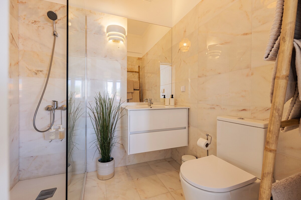 A modern bathroom is displayed with light-colored marble walls. A transparent shower enclosure is visible alongside a sleek, rectangular vanity with a mirror above. A potted plant adds a touch of greenery, while a rolled towel is neatly arranged on a wooden rack.