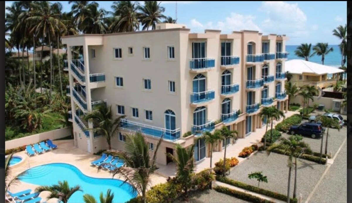 A modern three-story building with a light-colored facade is depicted, featuring multiple balconies with blue railings. In the foreground, a clear swimming pool is surrounded by lounge chairs and landscaped gardens, with palm trees visible throughout the area.