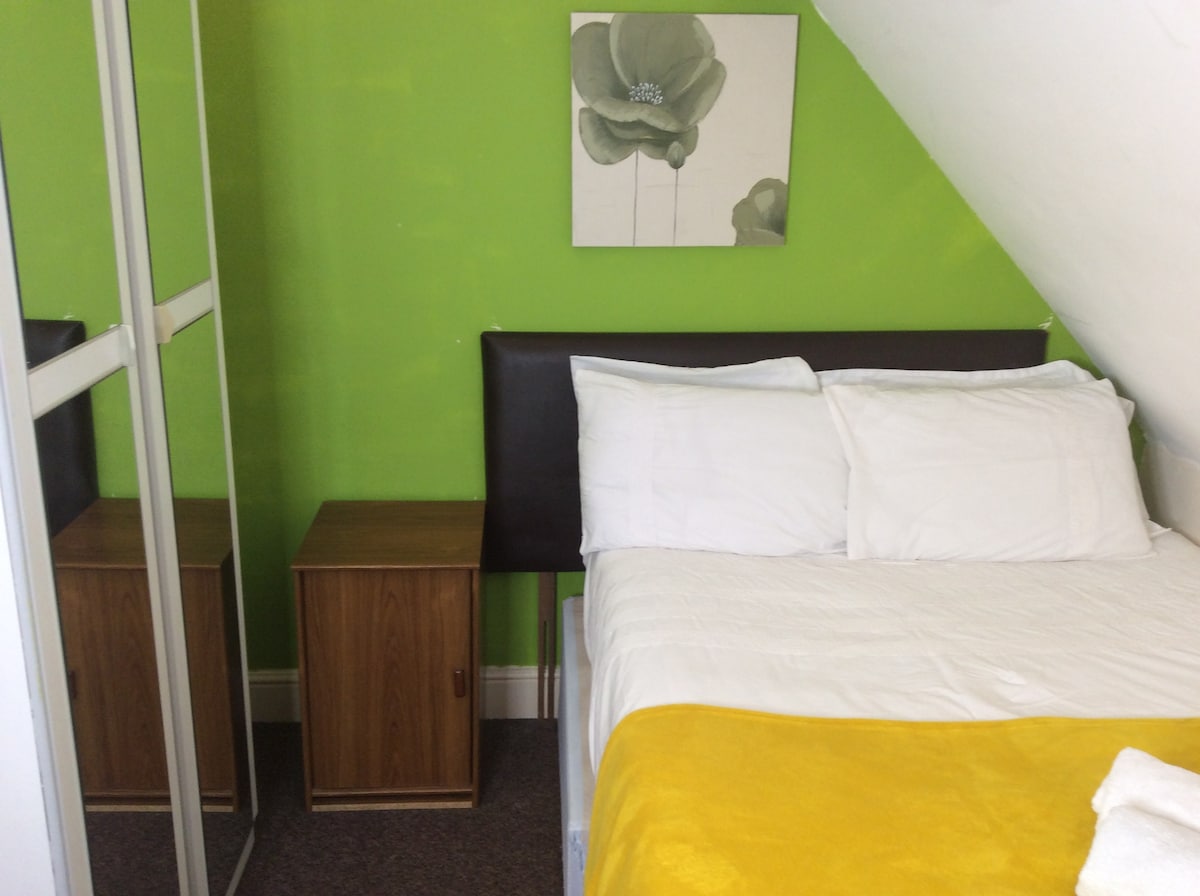 A cozy bedroom features vibrant green walls, a large bed with white linens, and a yellow blanket at the foot. There are two wooden nightstands alongside the bed, and a mirrored wardrobe reflects the room's details.