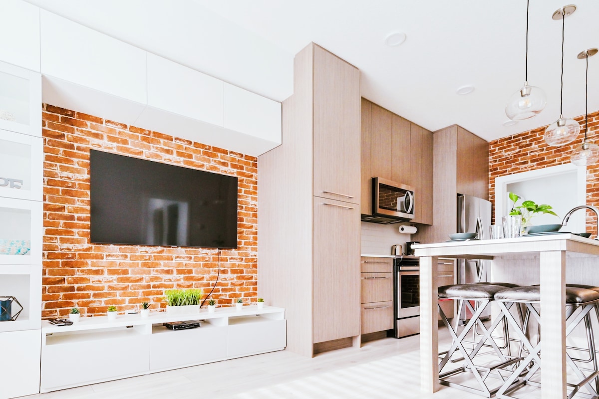 A modern kitchen space features a stylish brick accent wall and sleek cabinetry. Stainless steel appliances are positioned for convenience, while a dining table with metal chairs offers space for meals. Natural light streams in, creating a bright and inviting atmosphere.
