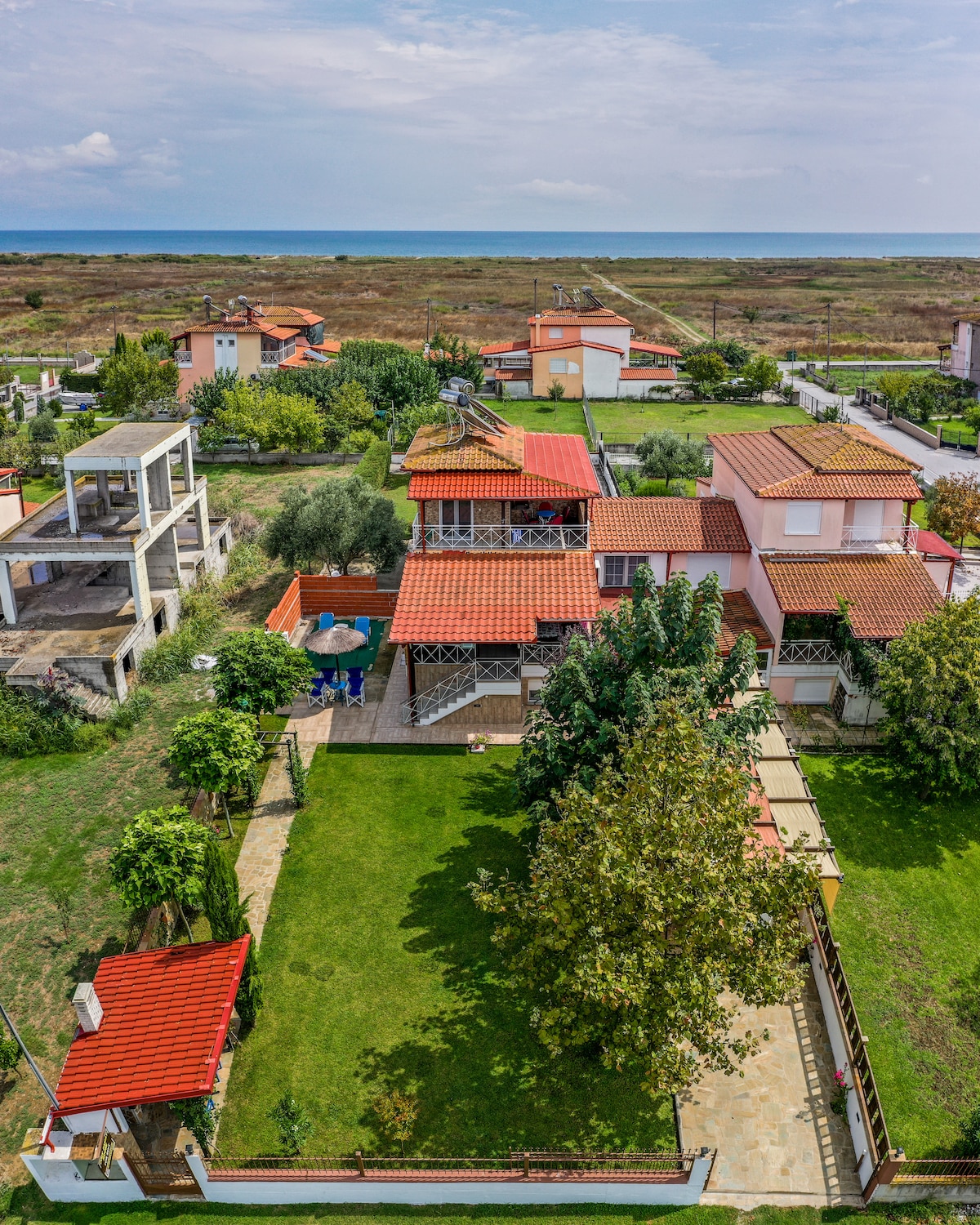 An aerial view showcases a spacious yard with lush green grass and mature trees. A multi-level house with a terracotta roof is nestled within the landscape, with nearby residential buildings visible in the background. The coastline can be seen in the distance, highlighting the serene environment.