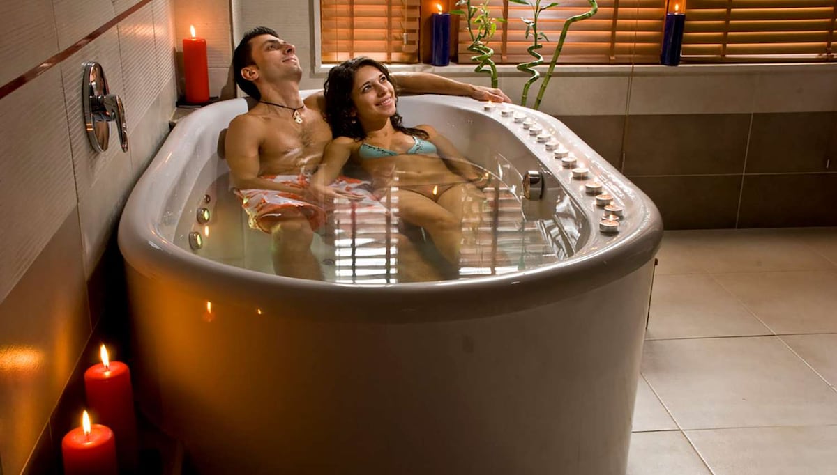 A spacious bathtub is filled with water and surrounded by candles, creating a relaxed ambiance. Nearby, natural light filters through wooden blinds, highlighting the soothing environment. Decorative elements such as plants add a touch of greenery to the scene.