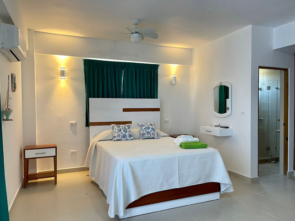 A spacious bedroom is equipped with a large bed, complemented by decorative pillows. A ceiling fan provides airflow, and natural light filters in from a window covered with teal curtains. A mirror and bedside tables enhance the functionality of the room.