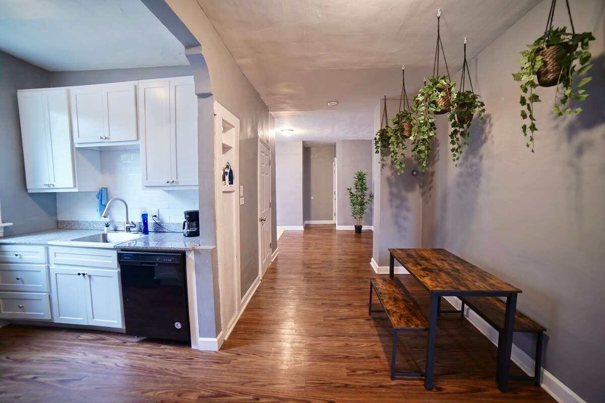 A bright corridor leads to a kitchen featuring white cabinetry and dark appliances, including a dishwasher. A rustic wooden dining table with metal legs is positioned nearby. Lush green plants hang from the ceiling, bringing a touch of nature into the space.