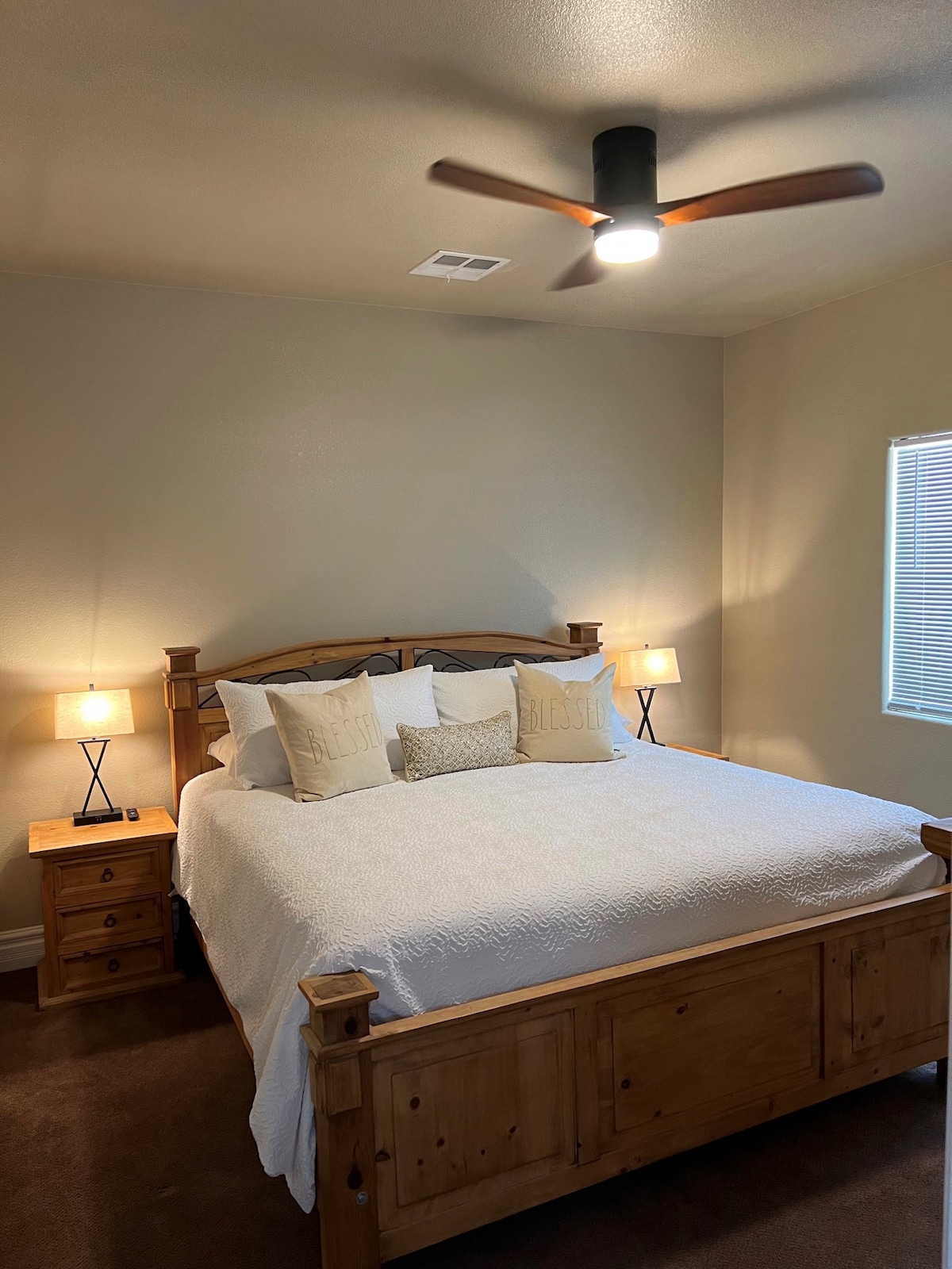 A spacious bedroom features a wooden bed frame with a large white comforter. Two bedside tables with lamps are positioned on either side. A ceiling fan hangs above, and natural light enters through a window dressed with blinds.