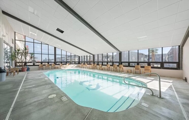 An indoor heated pool is situated in a spacious area, surrounded by large windows that provide ample natural light. Chairs are arranged along the walls, and lush plants add a touch of greenery to the serene environment.