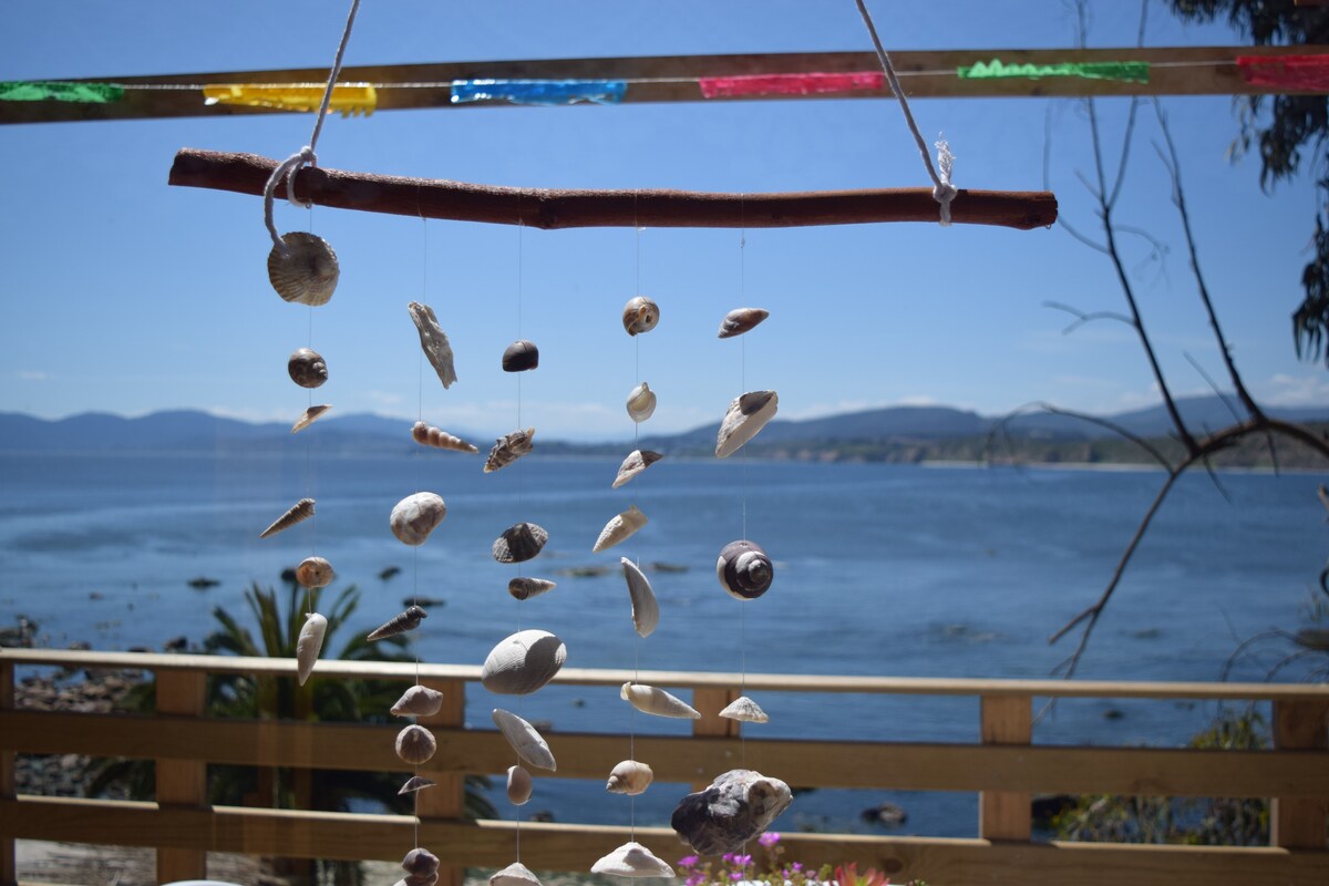 A shell mobile hangs from a wooden rod, decorated with colorful flags. The mobile features various sea shells, swaying gently in the breeze, with a clear view of the ocean and distant hills in the background.