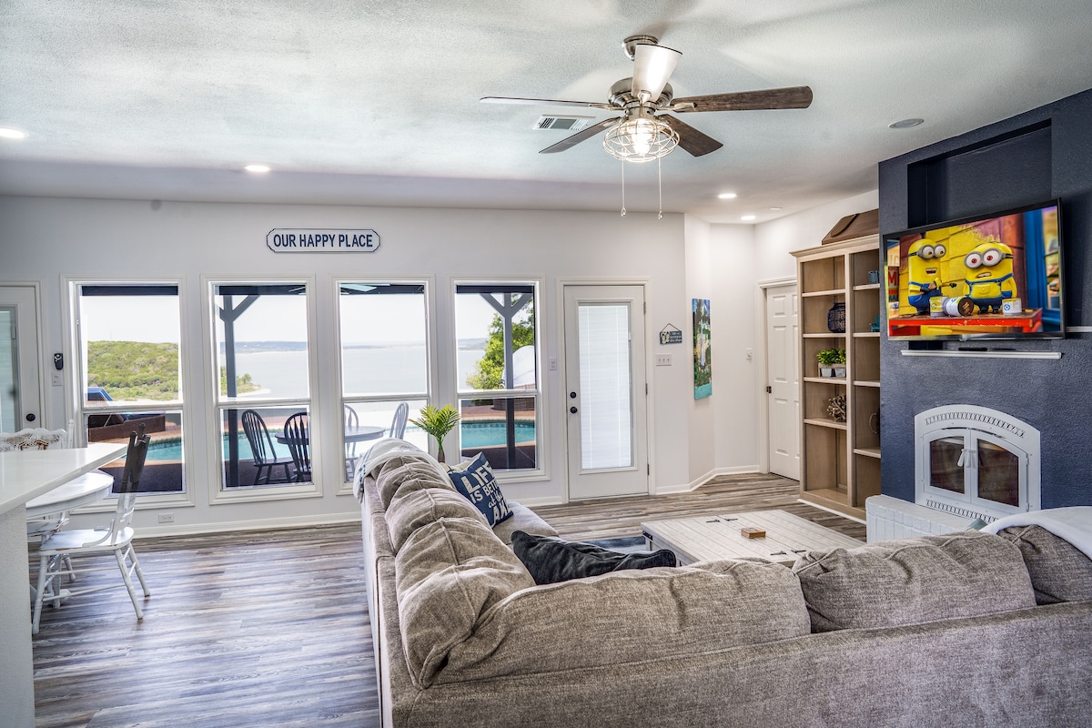 A spacious living area features a large L-shaped sectional sofa facing a wall-mounted television. Large windows provide a view of the lake and an outdoor dining area, while a ceiling fan adds to the comfortable atmosphere.