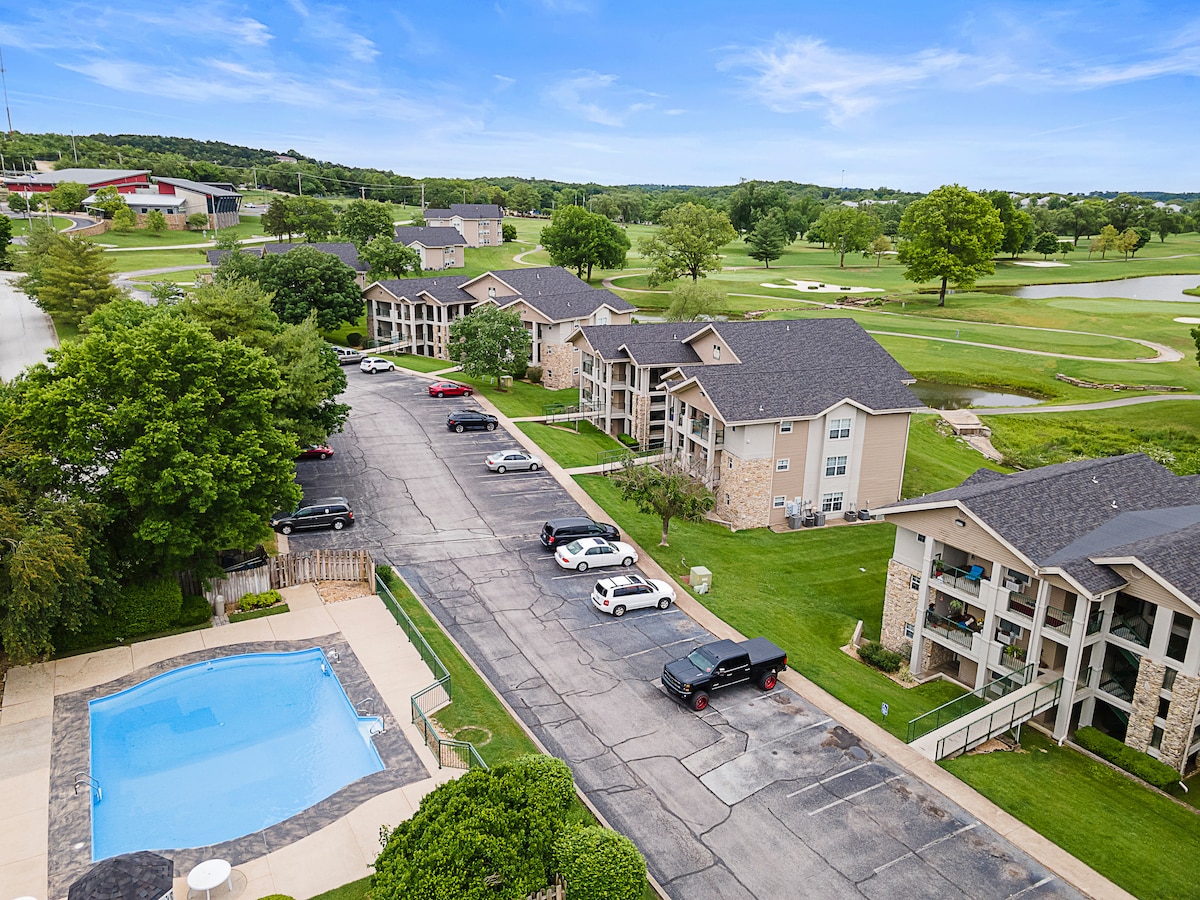 An aerial view showcases the Holiday Hills Resort, featuring well-maintained buildings that surround a lawn area and golf course. A seasonal outdoor pool is visible in the foreground, along with a parking lot accommodating various vehicles, all set against a backdrop of green landscaping.