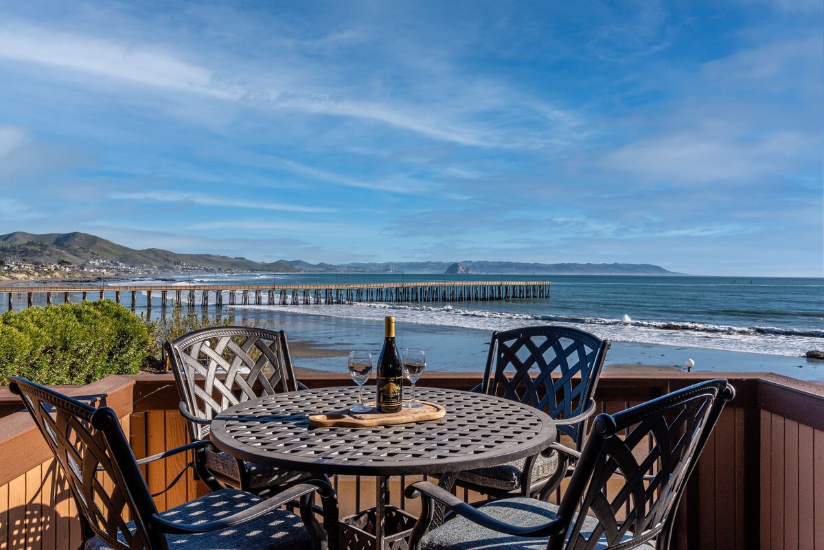 A round outdoor dining table is set with two glasses and a bottle of wine. The backdrop features a view of a long pier extending into the ocean, with gentle waves lapping at the shore under a bright blue sky.