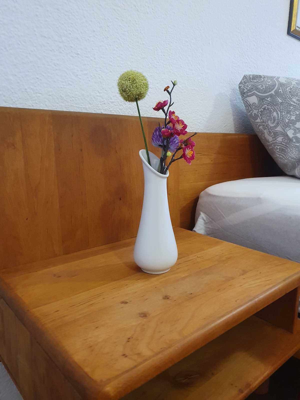 A white vase is positioned on a wooden nightstand, showcasing vibrant flowers in shades of pink and green. The nightstand features a clean design, complementing the nearby upholstered bedding that is partially visible.
