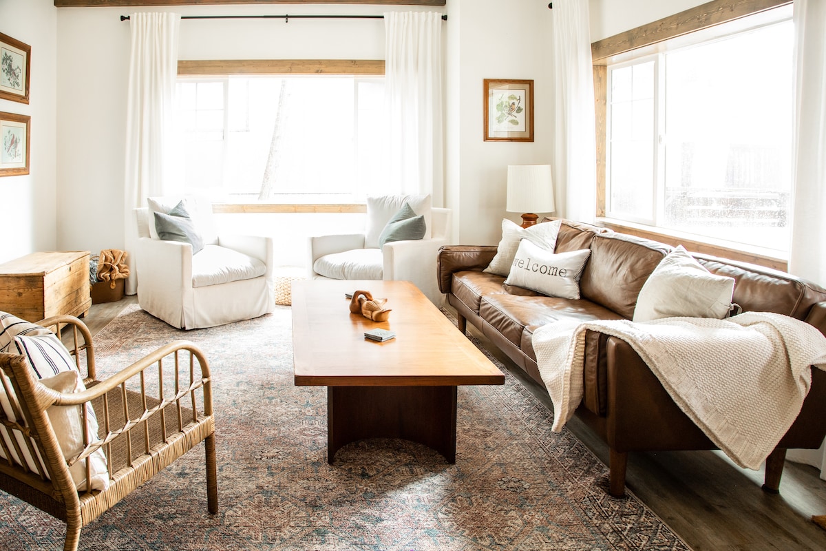 A spacious living area is designed with a brown leather sofa paired with light-colored armchairs. Natural light fills the room through large windows adorned with white drapes. A wooden coffee table sits at the center, complemented by a decorative throw and a cozy rug beneath.
