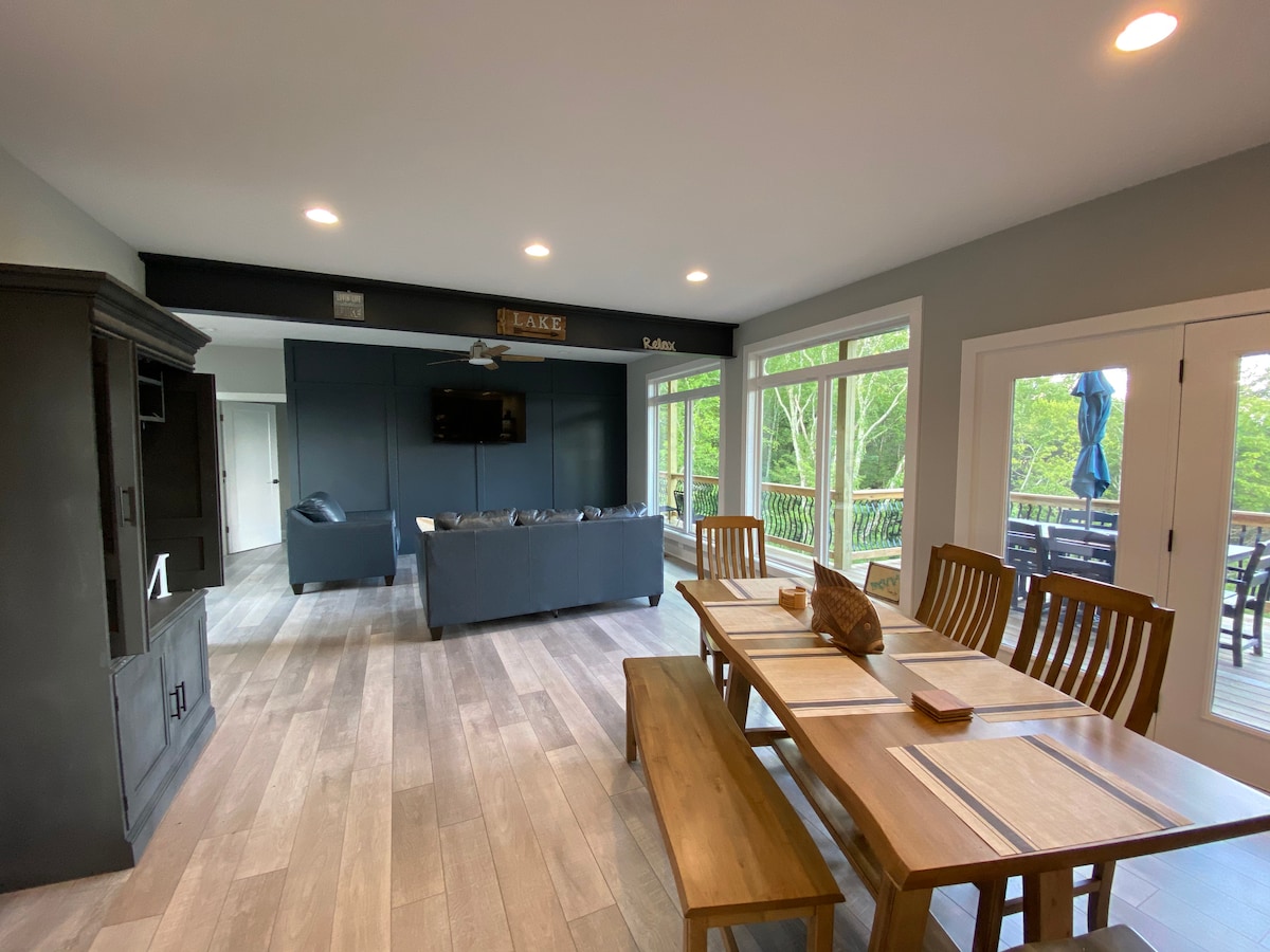 An open concept living and dining area features a large wooden table, comfortably seating several. A cozy seating arrangement is visible, facing a mounted television against a dark wall. Large glass doors provide access to the back deck, showcasing views of the surrounding greenery.