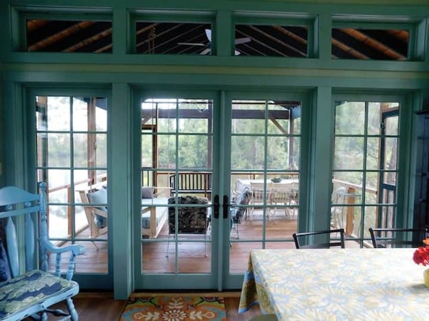 Double doors framed in soft green paint open to a spacious outdoor deck, revealing views of a serene pond surrounded by trees. A bright tablecloth covers the dining table, and a cozy seating area can be seen on the porch.