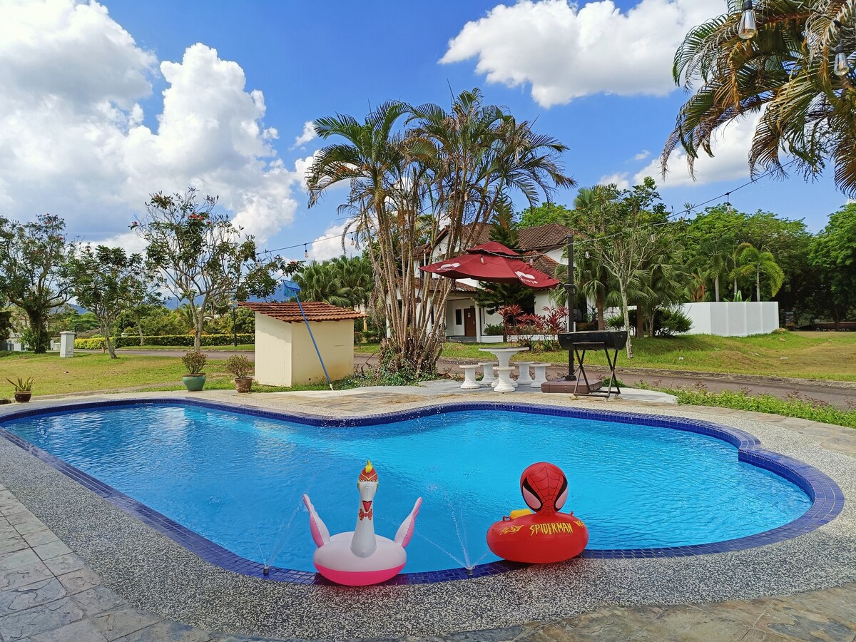 The private swimming pool is surrounded by well-maintained landscaping. A patio table and chairs are situated nearby, offering a shaded area under an umbrella. Colorful inflatable floats are positioned in the pool, contrasting with the clear blue water and the vibrant greenery in the background.