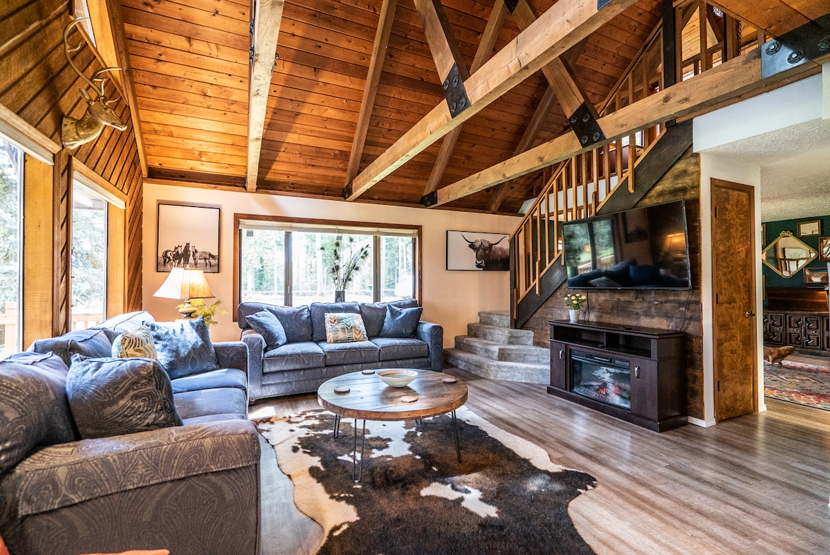 An open living area features a high, wooden beamed ceiling and large windows allowing natural light to filter in. A comfortable seating arrangement is positioned around a circular coffee table, while a flat-screen television is mounted on the wall. Light hardwood flooring complements the rustic aesthetic.