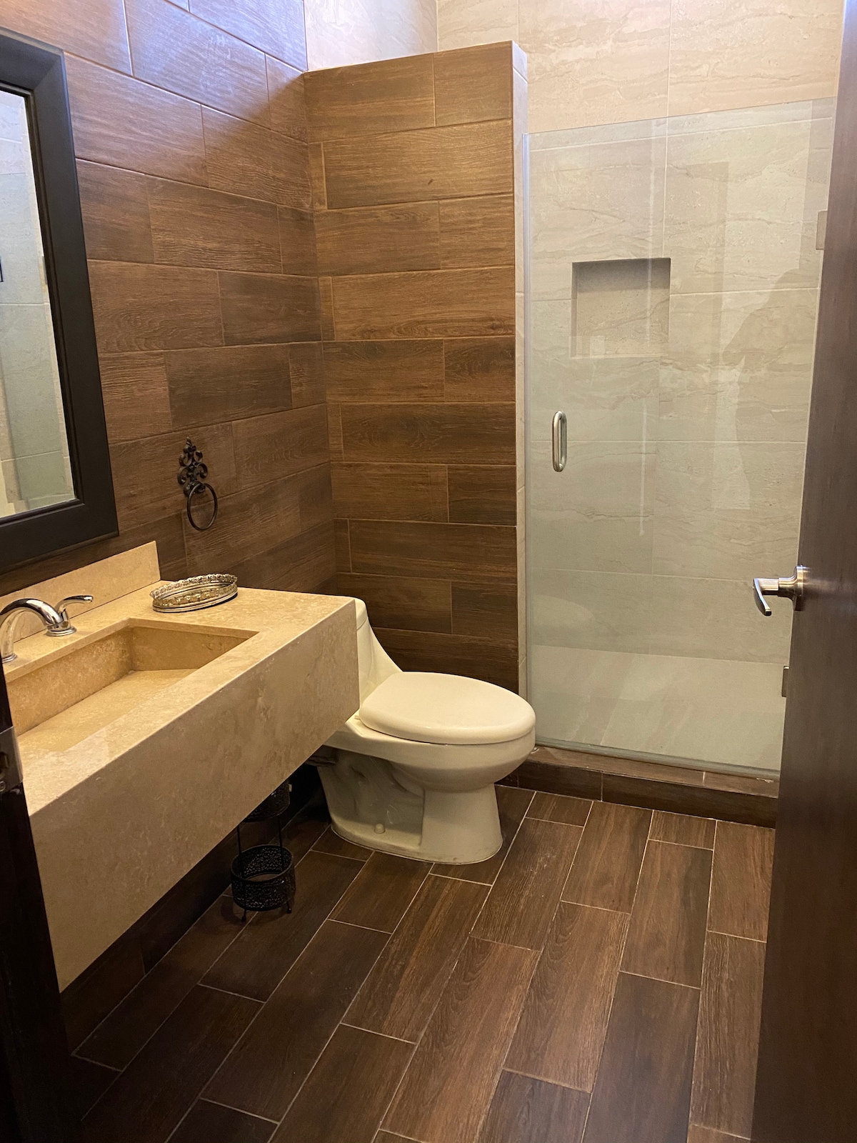 A modern bathroom is shown featuring wooden wall tiles and a glass shower enclosure. A stone sink is positioned beside a toilet, and a small decorative item rests on the counter. Natural light enters through the frosted window, enhancing the warm tones of the space.