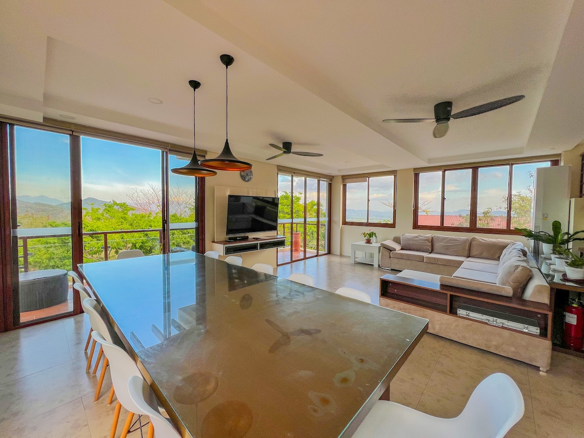 A spacious living and dining area is presented, featuring large windows that offer views of the surrounding mountains. The room includes a long dining table with white chairs and a comfortable sectional sofa. A television is mounted on the wall, and ceiling fans are visible.