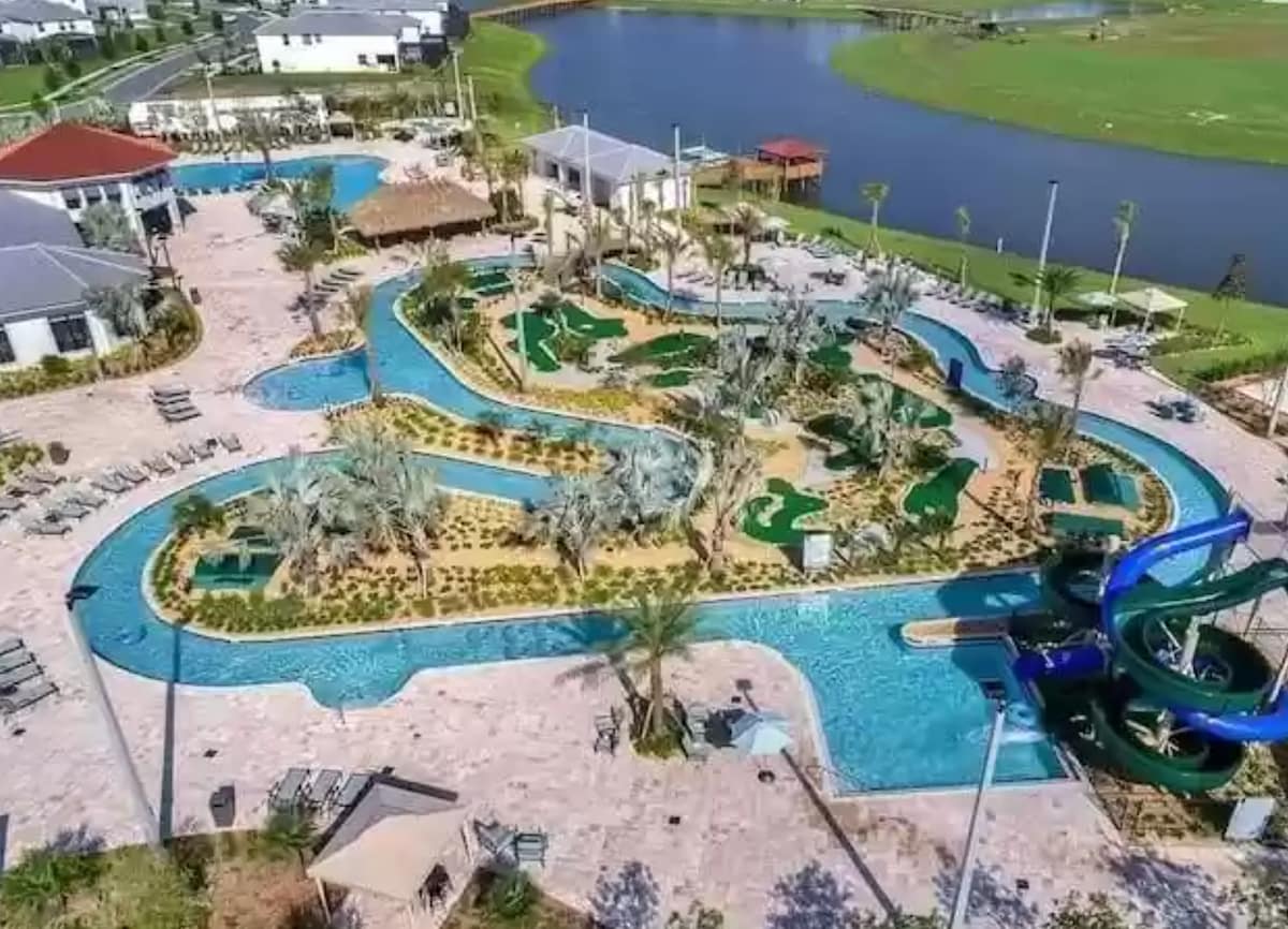 A sprawling resort pool area is displayed, featuring winding water slides, a lazy river, and lush landscaping with palm trees. Sun loungers line the pool, while shaded cabanas provide relaxation spots. Nearby, green spaces are visible, enhancing the recreational setting.