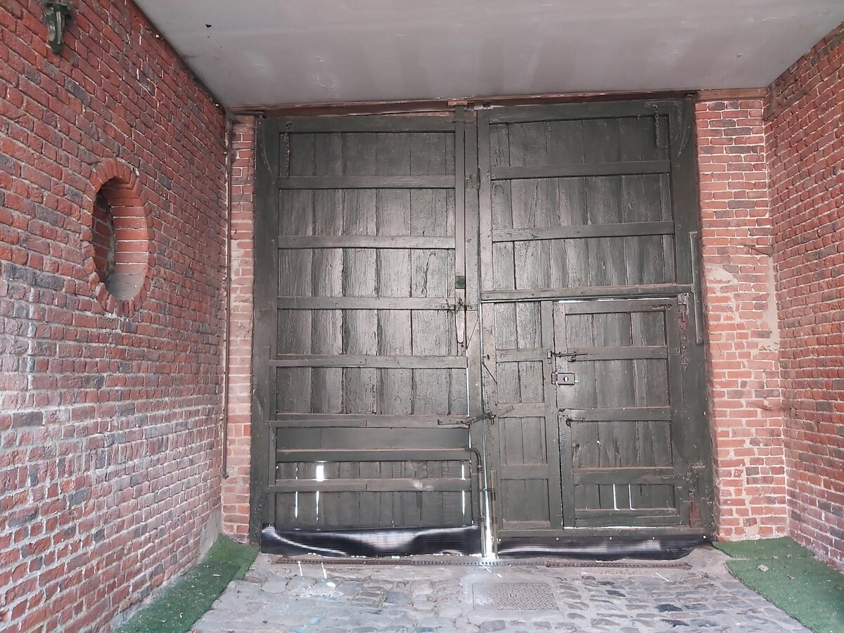 A pair of large, rustic green wooden doors stands closed, framed by exposed brick walls. The textured surface of the doors contrasts with the smoothness of the surrounding brick. A circular opening is visible in one wall, allowing a glimpse of the exterior.