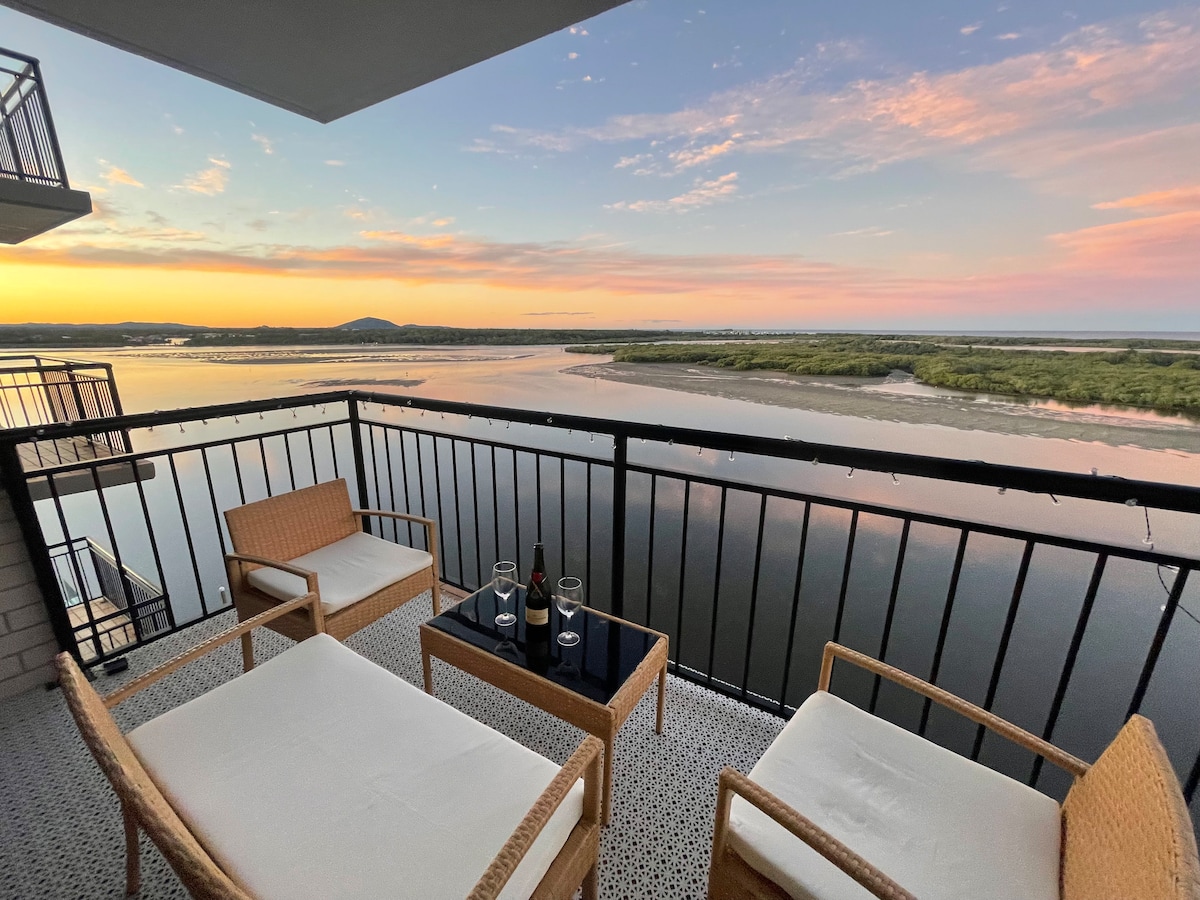 An expansive balcony offers panoramic views of the river and coastal landscape during dawn. Two wicker chairs flank a small table, providing a serene space for relaxation, while soft hues of pink and orange illuminate the sky, reflecting on the water.