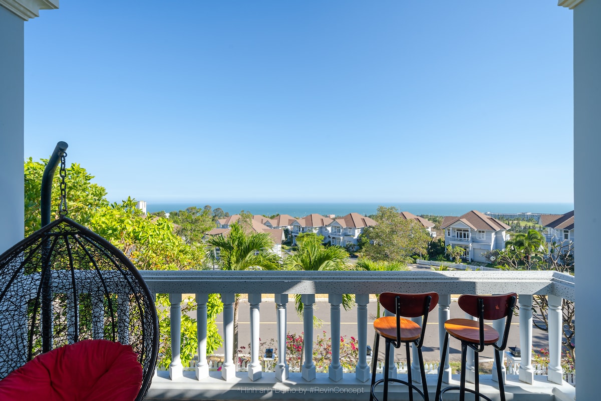 A balcony view showcases a horizon meeting the ocean, framed by residential rooftops and lush greenery beneath a clear blue sky. Two bar stools and a hanging chair provide comfortable seating, inviting relaxation while enjoying the scenery.