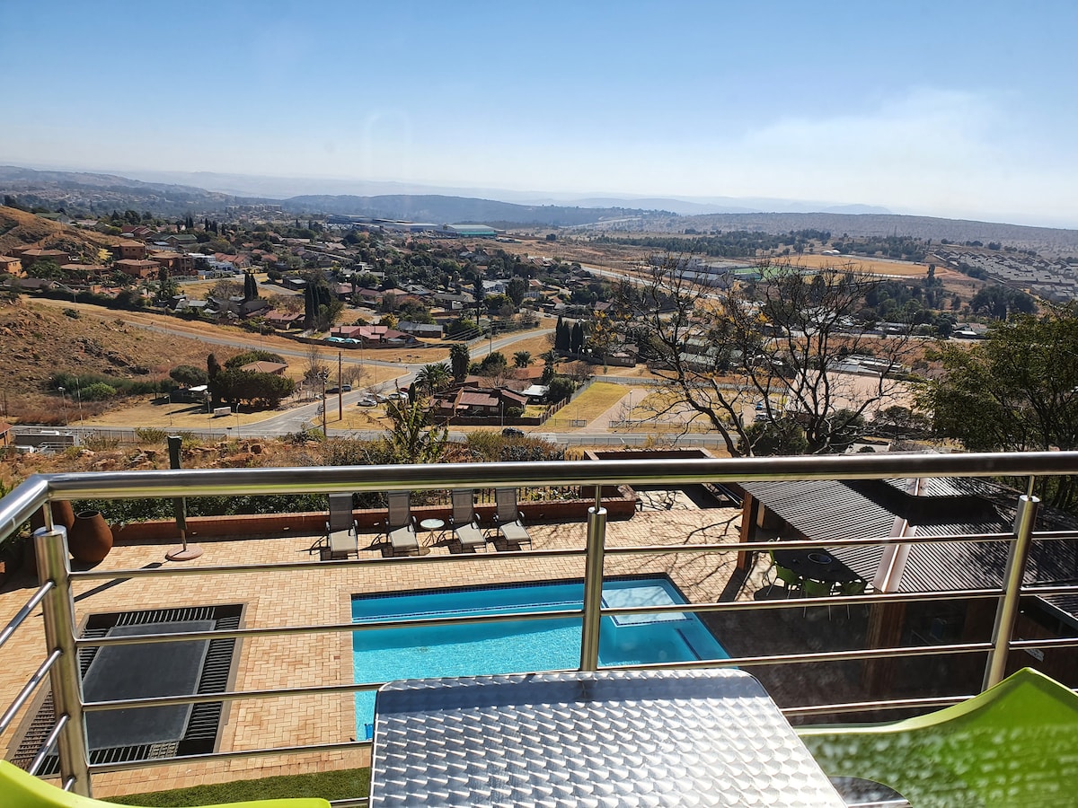 A vibrant view from the balcony features the expansive Magaliesburg landscape. The scene includes a clear blue sky, distant mountains, and a glimmering swimming pool below. Modern outdoor furniture is positioned on the balcony, inviting guests to relax and enjoy the serene surroundings.