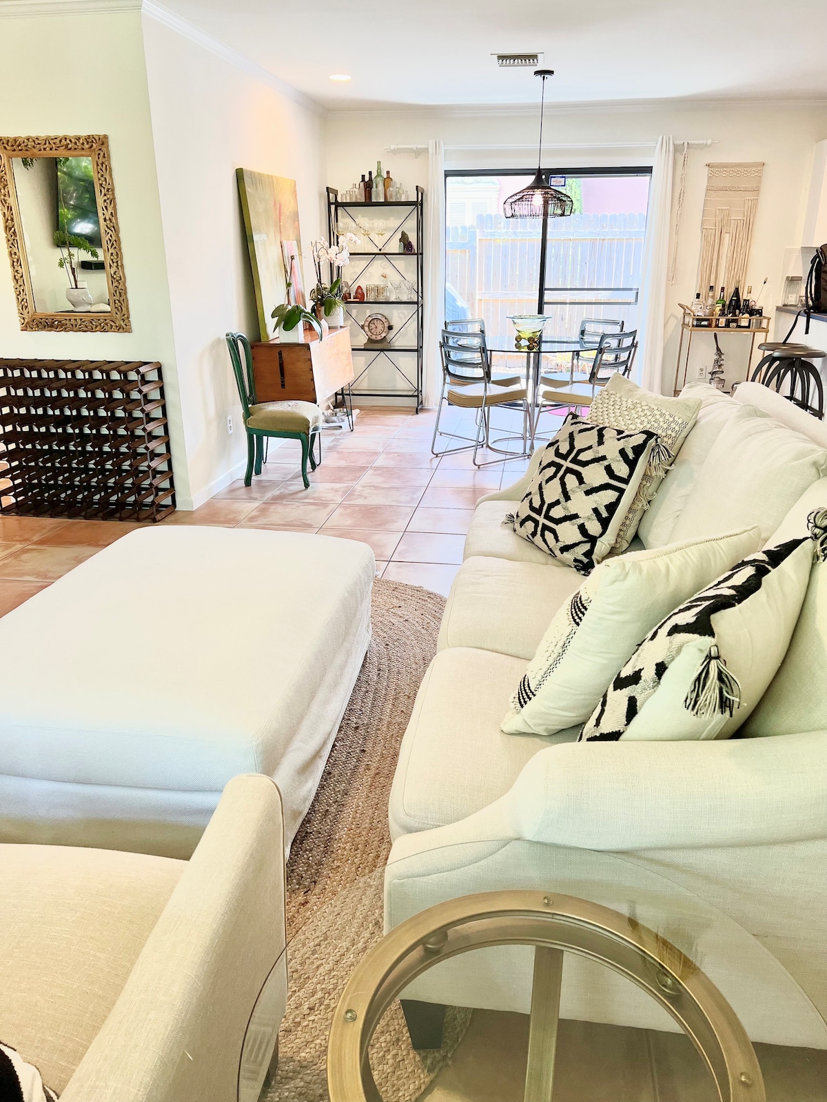 An inviting living area features a large white sofa adorned with patterned throw pillows. A round coffee table complements the seating arrangement. The open layout allows for a view of a dining space with a glass-topped table and six metal chairs, illuminated by natural light from large sliding doors.