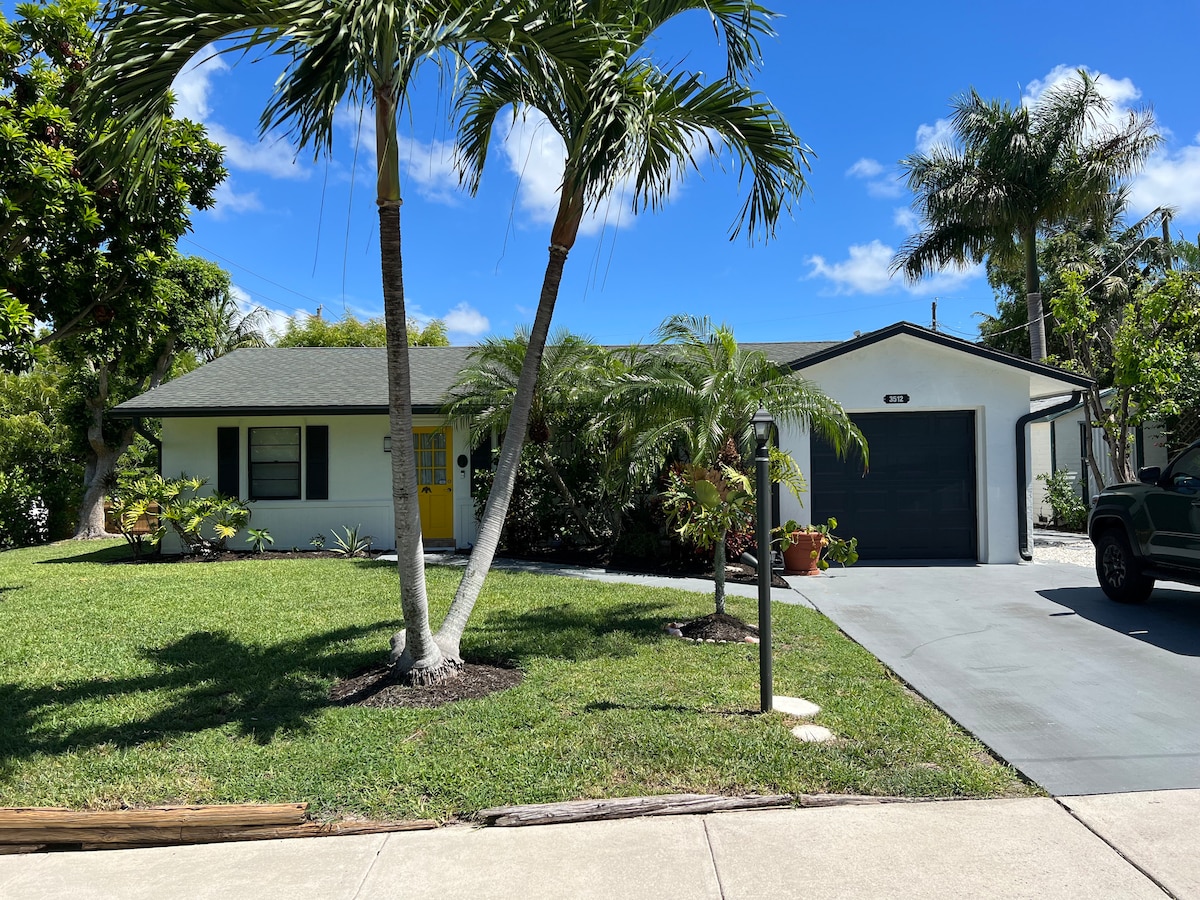 A single-family home is depicted with a well-maintained front yard featuring tropical landscaping and mature palm trees. The house is painted in light colors, complemented by a bright yellow door. A concrete driveway leads to the garage on the right side.