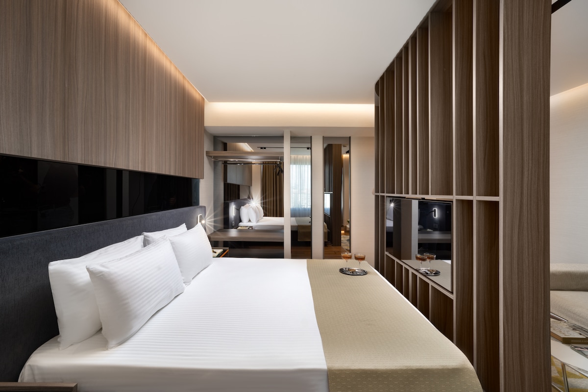 A spacious sleeping area features a neatly made bed with crisp white linens and a textured beige throw. A decorative separator divides the lounging space, which includes a mounted television. Natural light flows in through a large window, complemented by warm wooden accents throughout the room.