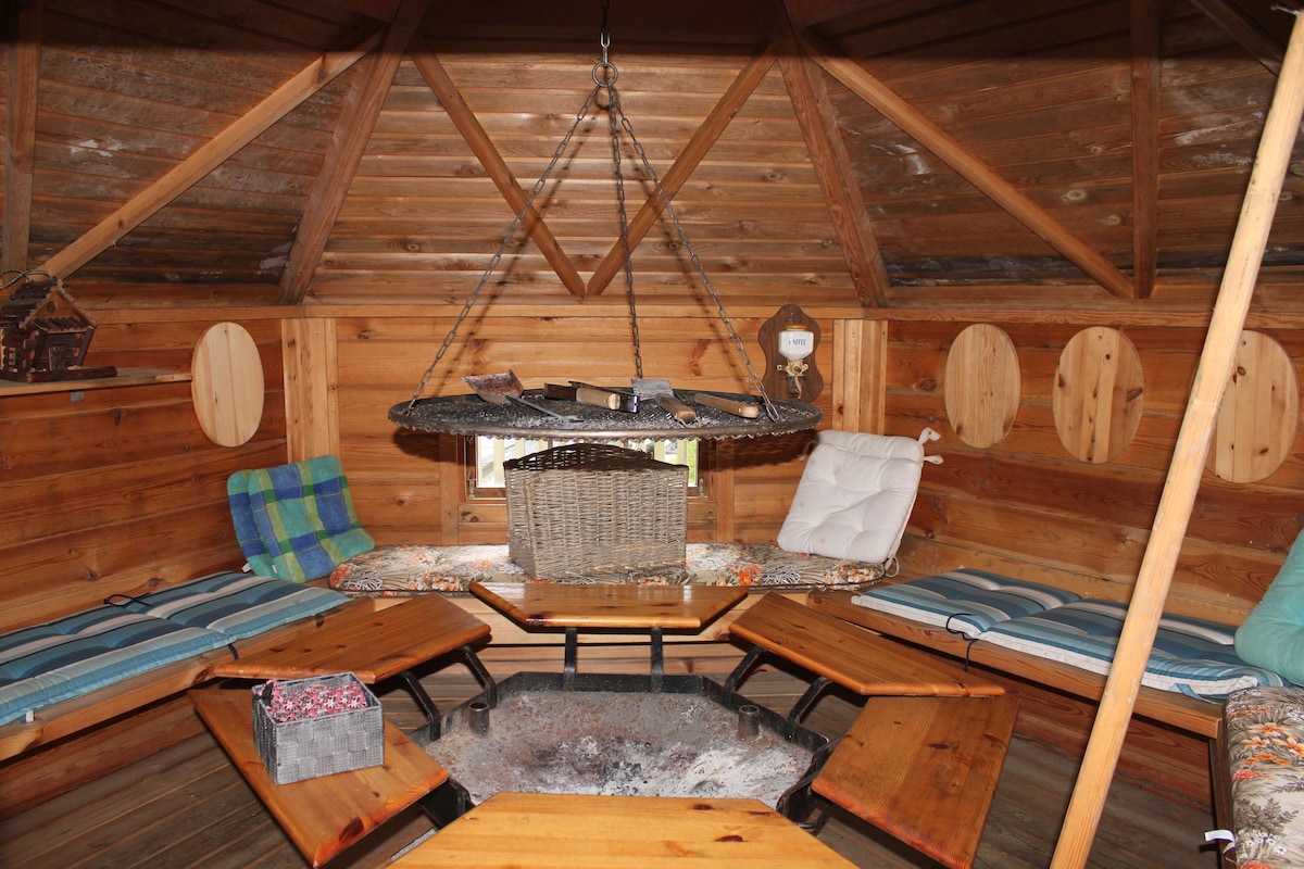An inviting grill hut features wooden walls and a high, angled ceiling. A round, suspended grill hangs above a fire pit surrounded by several sturdy wooden tables. Cushioned seating lines the walls, offering comfort for gatherings. Natural light filters in through windows.