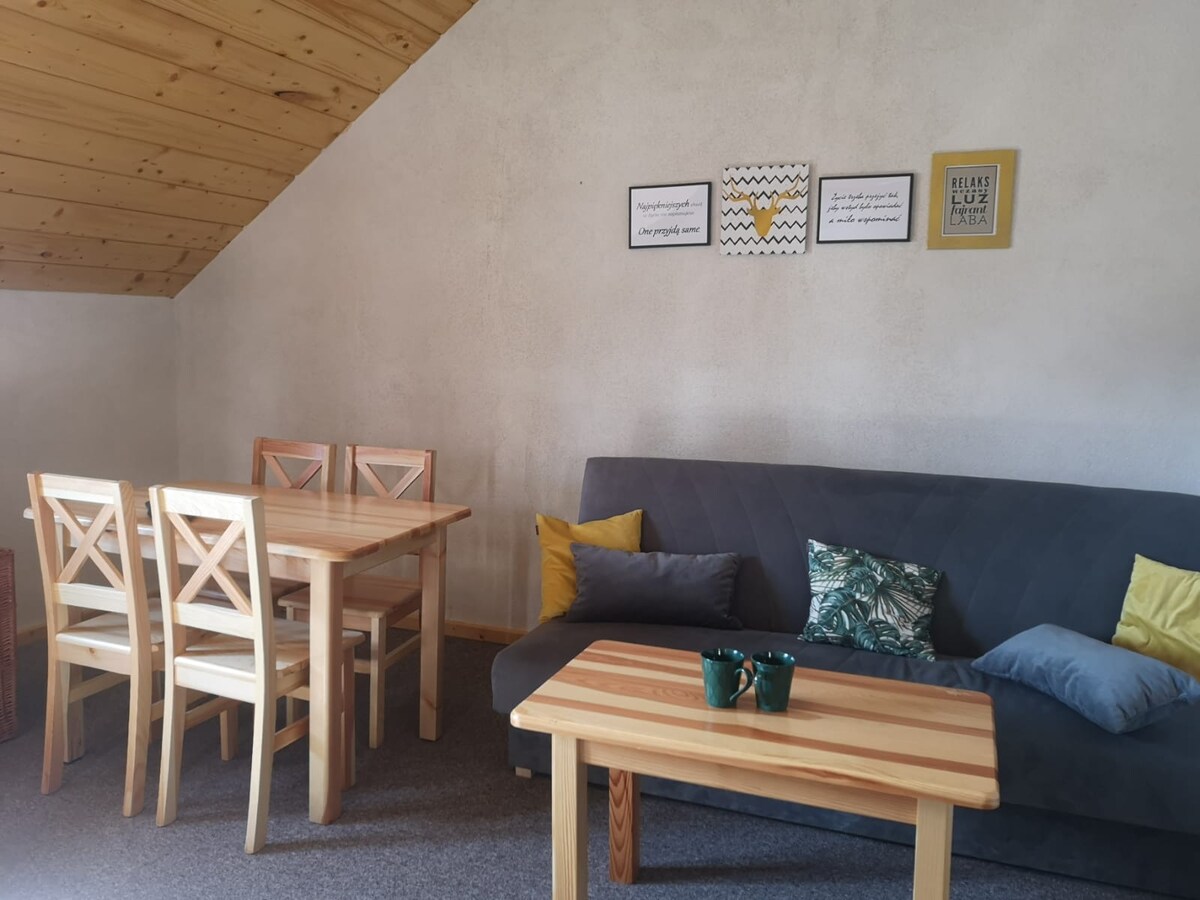 A cozy seating area features a dark gray sofa adorned with colorful throw pillows. A wooden coffee table sits in front, complemented by a dining table with four wooden chairs. Wall decor with inspirational quotes adds a personal touch to the space.