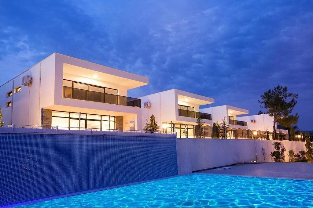 Three contemporary villas are positioned beside a tranquil pool, reflecting a calming blue. Each villa features large glass doors and private balconies that extend into the warm evening glow, surrounded by minimal landscaping and softly illuminated against a dusky sky.
