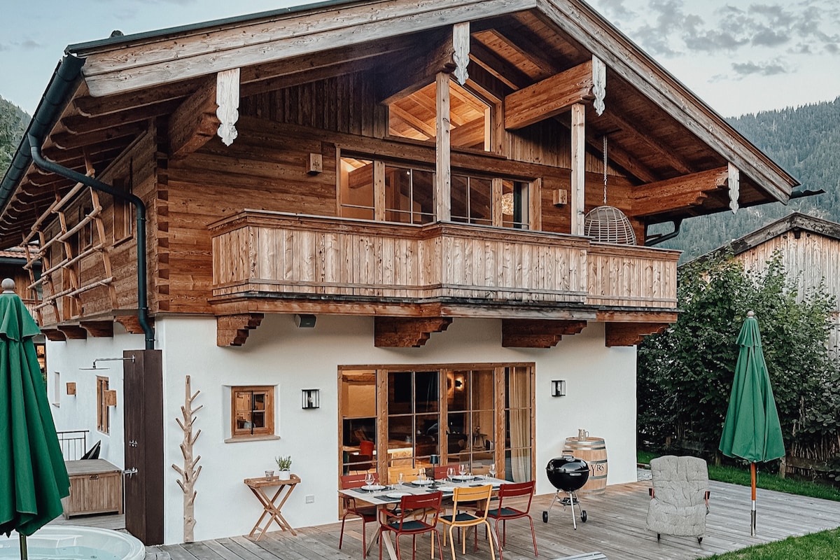 A charming chalet exterior is showcased, featuring a wooden balcony and expansive glass doors. Outdoor dining furniture is arranged on the wooden deck, accompanied by green umbrellas. The surrounding landscape includes lush trees and mountain views that enhance the serene setting.