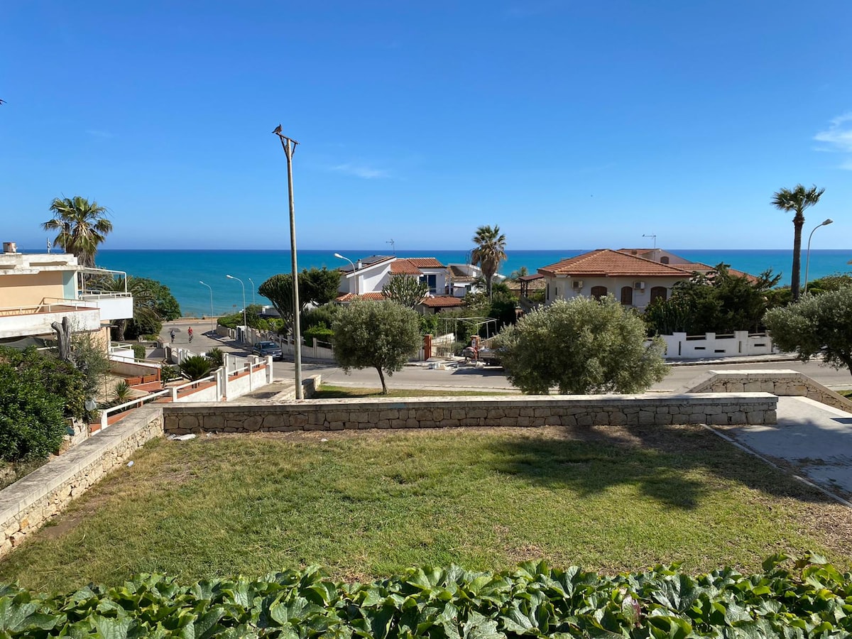 A clear view of the coastline is presented, showcasing the vibrant blue sea under a sunny sky. Flanking the foreground, a grassy area is bordered by gardens and palm trees. Residential buildings are visible in the background, contributing to a serene coastal environment.