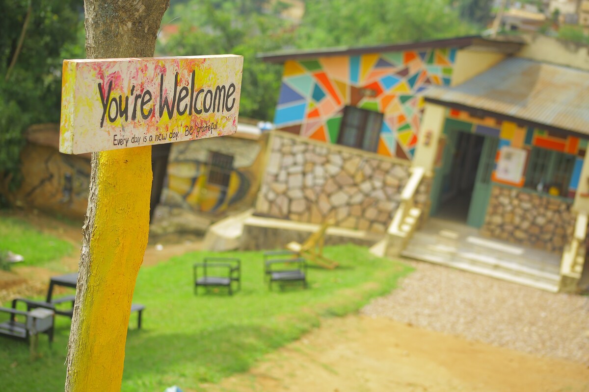 A colorful sign displaying 'You’re Welcome' is mounted on a tree, inviting guests. In the background, a vibrant mural decorates the exterior of a building, which features a combination of stone and painted surfaces. A grassy area with benches is visible in front of the structure.