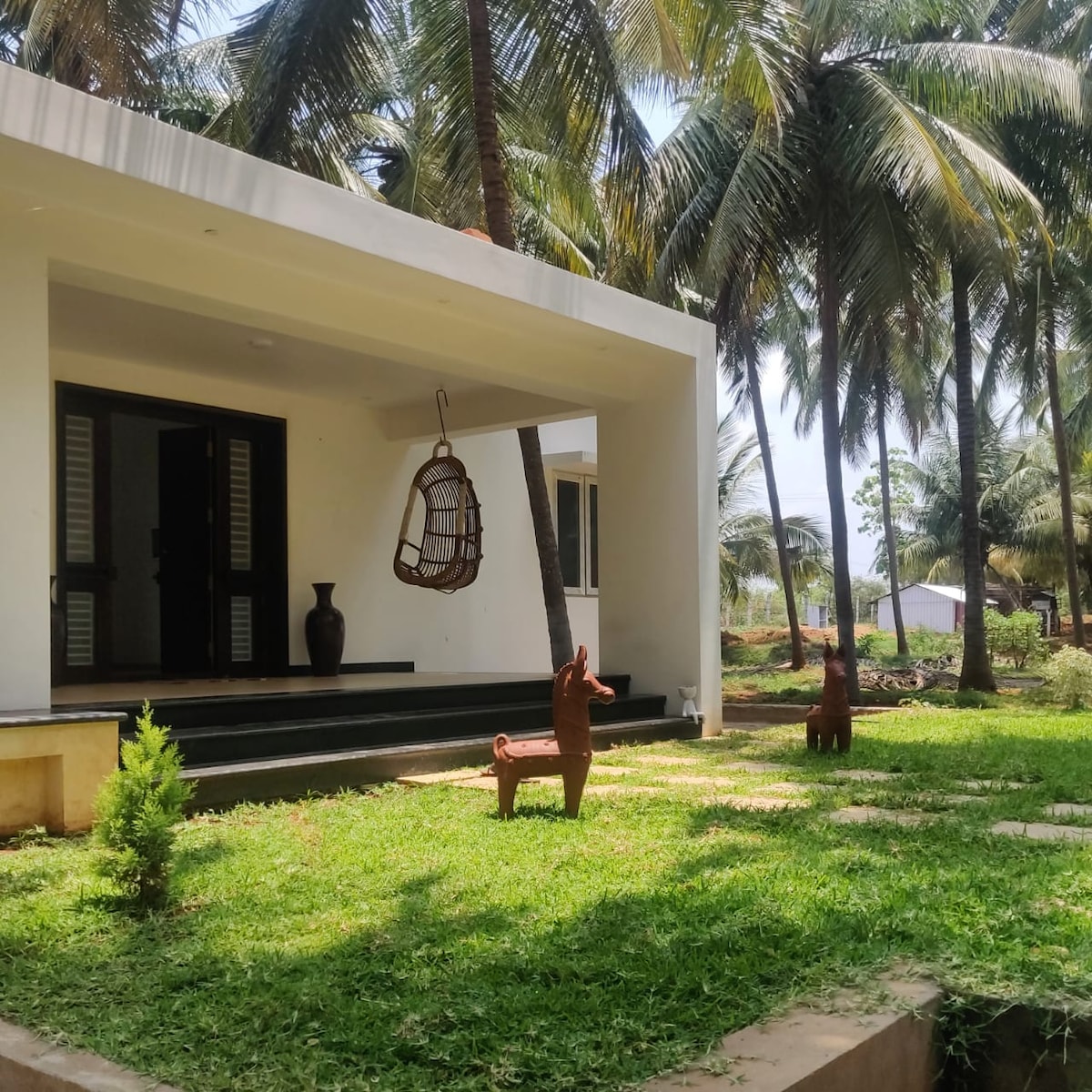 The entrance of the farmhouse features a well-maintained green lawn bordered by tall palm trees. A hanging chair is suspended from the porch ceiling, while two decorative animal sculptures are positioned on the grass, enhancing the welcoming outdoor space.