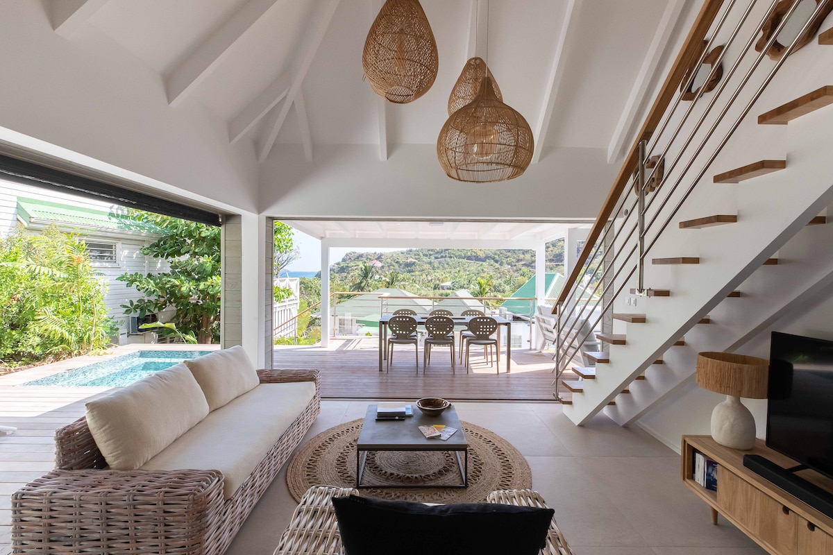 A modern living space features a light-colored sofa accentuated with a round area rug. A dining table with multiple chairs is visible, along with a staircase made of wood and metal. Large glass doors allow for views of the outdoors and a glimpse of the pool.