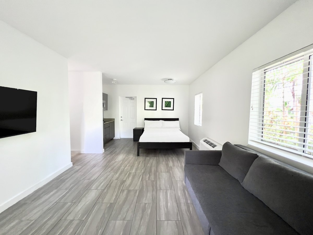 A spacious studio room features a queen bed with simple bedding. A comfortable sofa is positioned near a window with blinds. A flat-screen television is mounted on the wall, and a doorway leads to the kitchenette area. Neutral colors create a calming environment.
