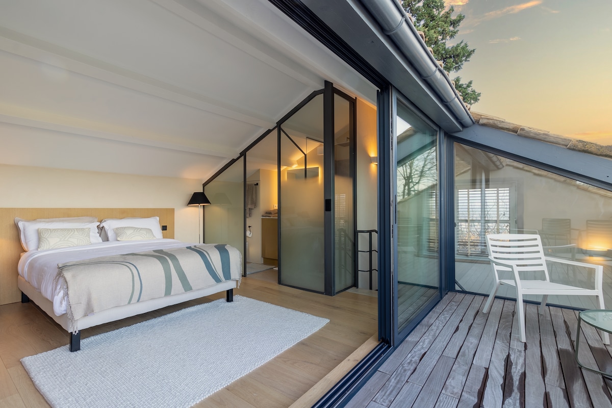A spacious bedroom features a comfortable bed dressed in neutral tones. Large glass doors open onto a wooden terrace, allowing natural light to fill the room. A cozy chair and table are positioned outside, providing an inviting space to enjoy the scenic views.