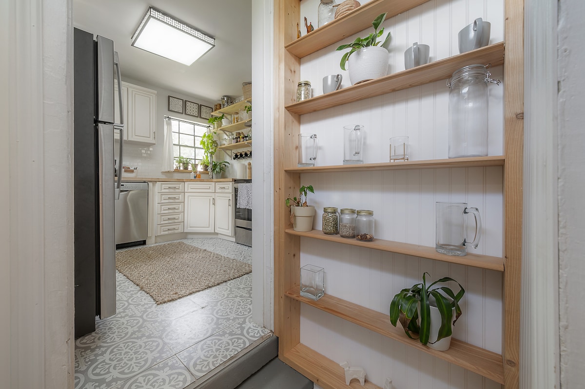 A well-organized kitchen space is visible through an open doorway, showcasing a fully stocked kitchen with shelves containing various jars and plants. A patterned tile floor complements the aesthetic. Natural light fills the space, enhancing the welcoming ambiance.