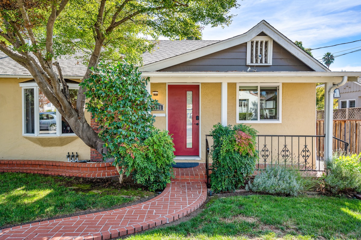 The home's exterior features a welcoming pathway made of brick, leading to a front door framed by a vibrant red color. Lush greenery and well-maintained landscaping surround the entrance, complemented by a shade-providing tree. The charming facade reflects a blend of architectural styles.