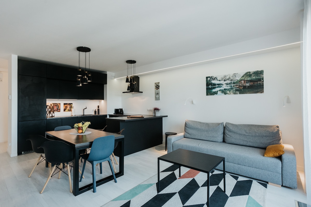A modern living area is presented with a neutral color palette. A spacious sofa is arranged alongside a dining table with four chairs. The kitchen features sleek cabinetry and is partially visible. Light fixtures hang above, creating a well-lit environment.