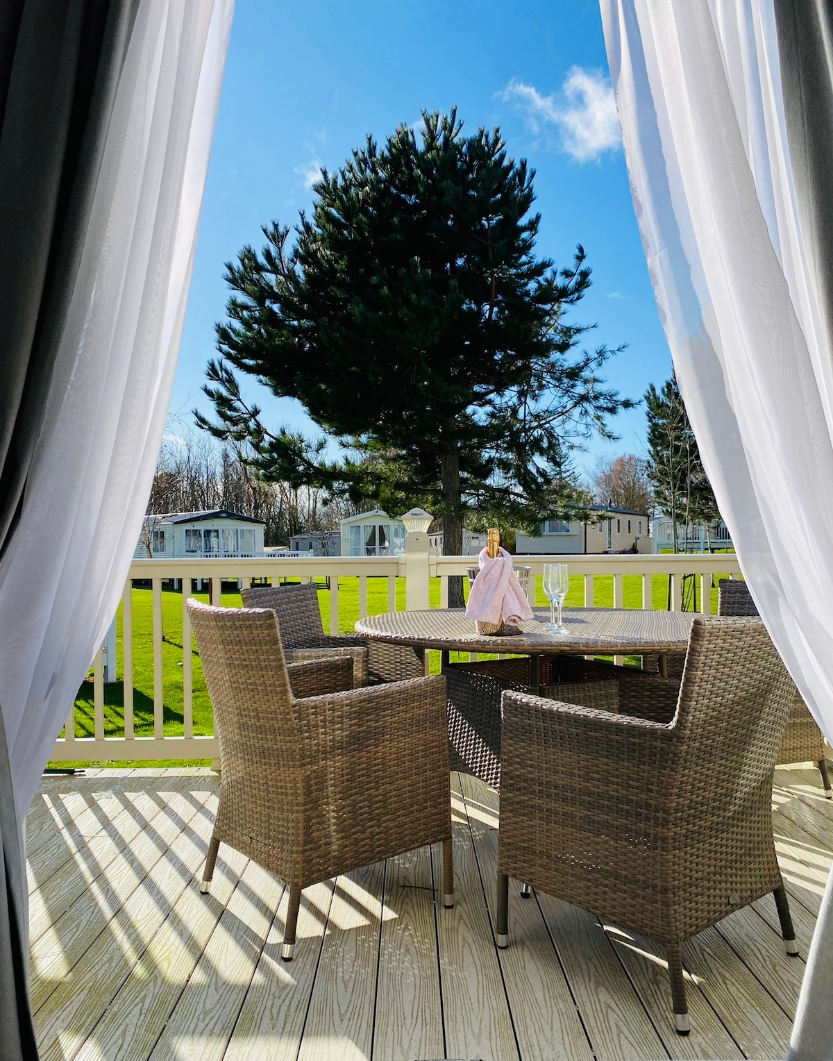 A patio area is visible through flowing sheer curtains, featuring a round table surrounded by woven chairs. A lone tree is seen in the distance, with rolling green grass and additional accommodations in the background under a clear blue sky.